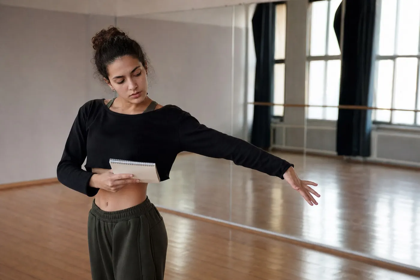 A young adult dancer alone in a sunlit empty studio, holding a notebook and marking choreography with her free arm, mirror and tall windows behind her